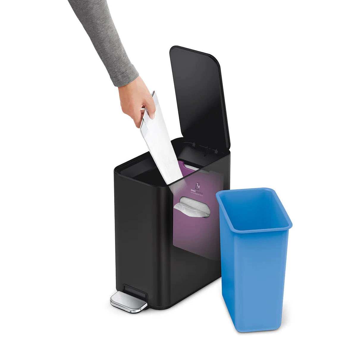 The image displays a black trash can with a rectangular design. The can features a lid being opened using a foot pedal, demonstrating its hands-free operation. Beside the trash can, there is a blue inner bin that is designed to fit inside it, enhancing ease of use and cleanliness by allowing easy removal and replacement of trash bags. A person's hand is shown disposing of a piece of paper into the trash can, which adds a dynamic element to the image and illustrates the functionality of the trash can in a re