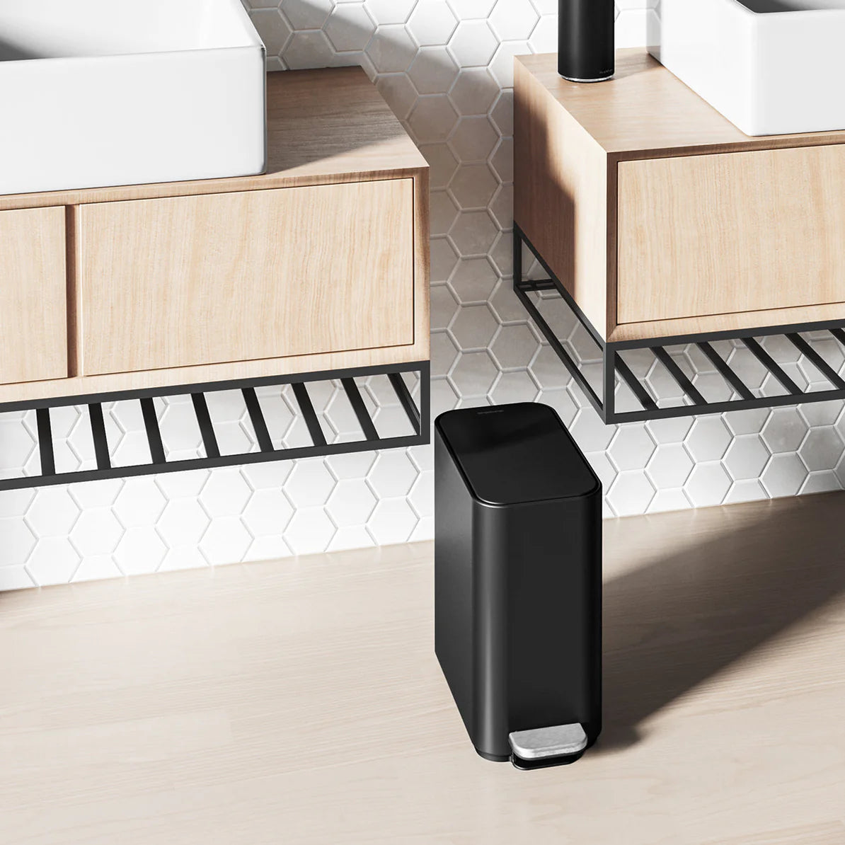 The image shows a black, rectangular trash can placed in a modern bathroom setting. The trash can is positioned on a light-colored floor between two floating wooden vanity cabinets, which have metal frames underneath. The bathroom features hexagonal white tiles on the wall, adding a contemporary touch to the space. The black trash can complements the minimalist and clean design of the bathroom, fitting neatly between the cabinets. The metallic foot pedal at the front adds a functional and stylish element, e