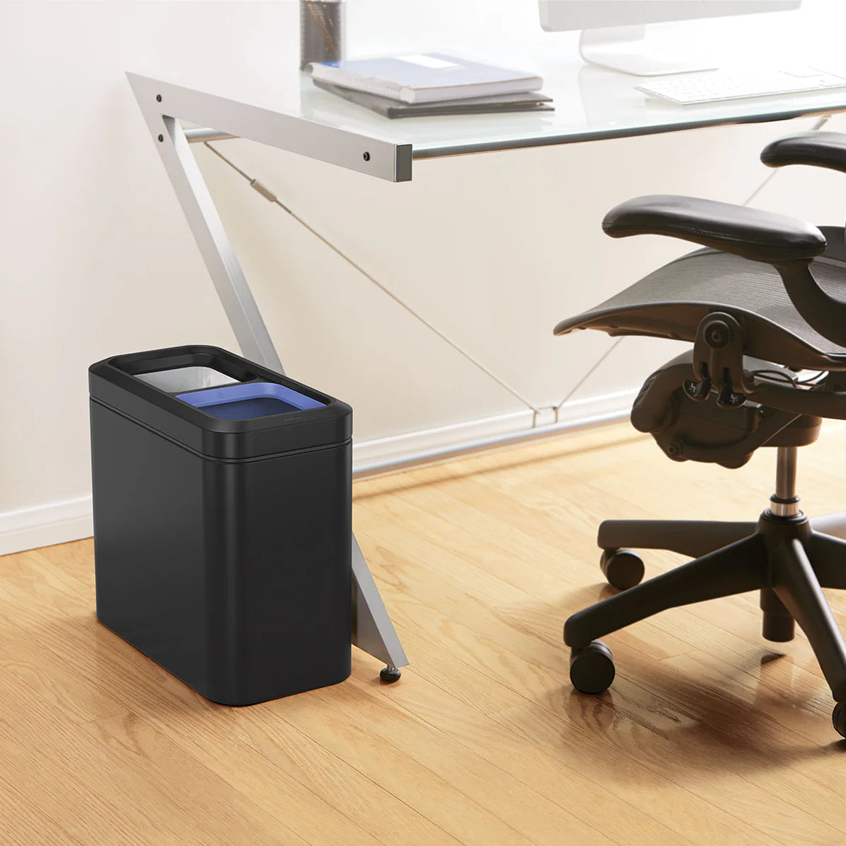 The image shows a modern office setting with a sleek, rectangular black trash can placed beside a glass-top desk. The trash can has a minimalist design with smooth, rounded edges and a matte finish, giving it a contemporary appearance. Inside the trash can, a blue liner is visible, indicating a dual-compartment or color-coded system. The compact size fits neatly next to the desk, making it convenient for waste disposal without taking up much space. The office chair, light wood flooring, and overall clean an