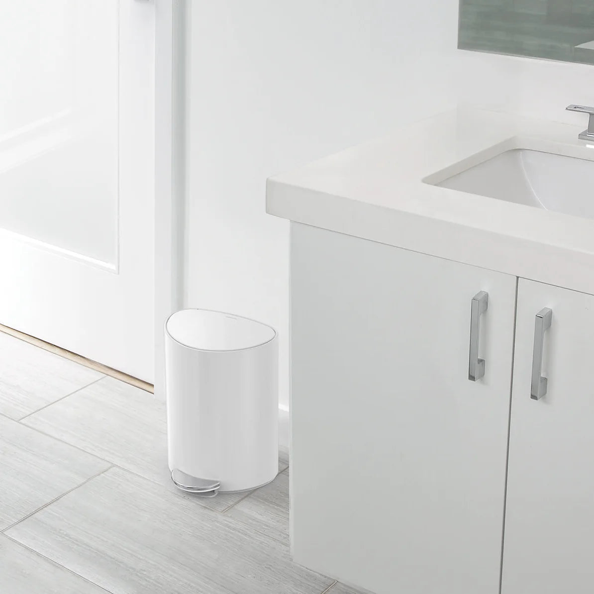 A clean, modern bathroom with light-colored tile flooring and white cabinetry. Next to the sink cabinet, there is a small, white, oval-shaped trash can with a sleek, minimalist design. The trash can has a polished chrome base and fits neatly into the space, blending seamlessly with the bathroom's bright and simple decor. The overall aesthetic is fresh, uncluttered, and contemporary.