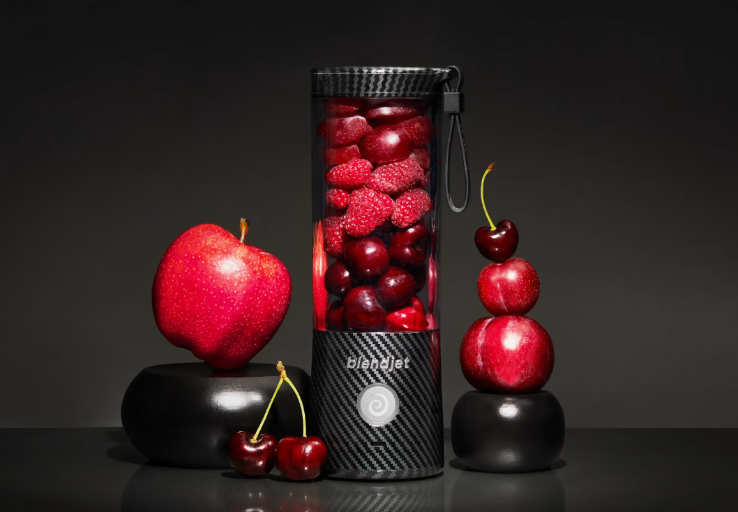 A carbon fiber-patterned BlendJet blender filled with raspberries and cherries, surrounded by a red apple, scattered cherries, and stacked fruits, set against a dark background.