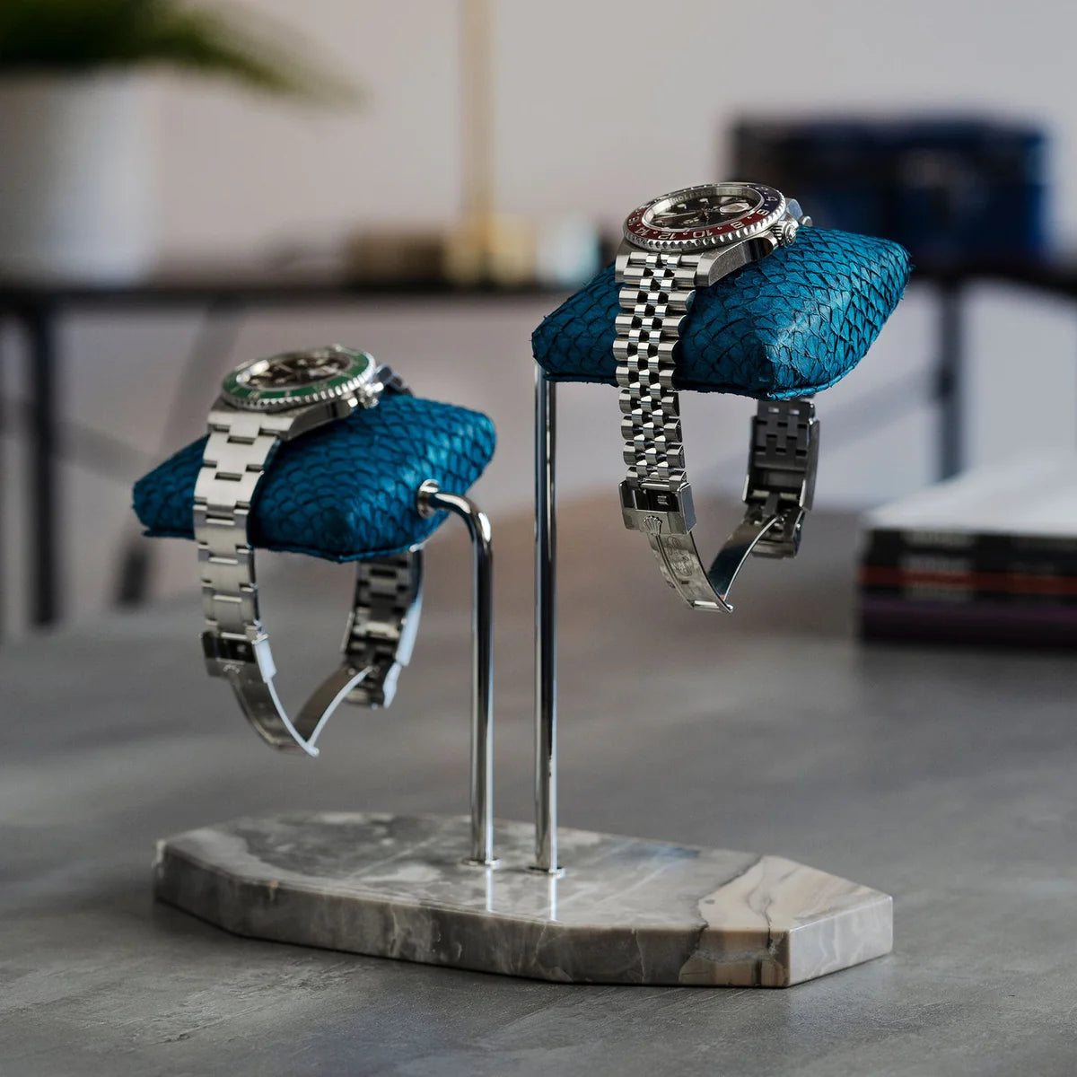  Two luxurious wristwatches are displayed on blue textured cushions, mounted on a double-arm stand with a gray marble base. One watch has a silver metal band with a black dial and green bezel, while the other has a silver metal band with a black dial and a red and blue bezel. The display is set on a gray surface, with a blurred background that includes a table, books, and a plant, adding to the sophisticated and stylish presentation.