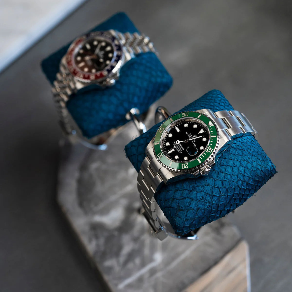 Two luxurious wristwatches are displayed on blue textured cushions, mounted on a double-arm stand with a gray marble base. One watch has a silver metal band with a black dial and green bezel, while the other has a silver metal band with a black dial and a red and blue bezel. The display is set on a dark surface, showcasing the elegant and sophisticated presentation of the watches.