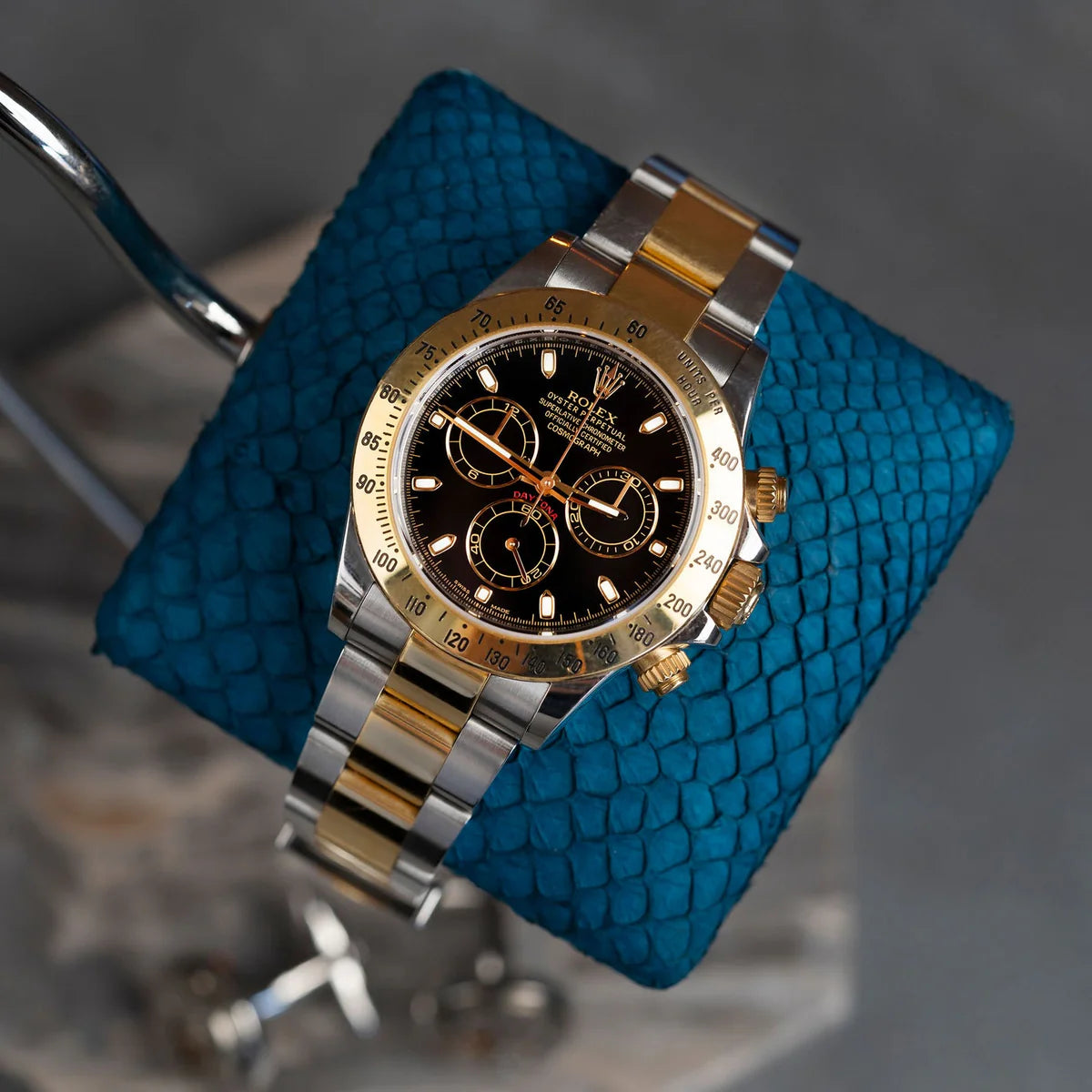 A luxurious wristwatch with a two-tone metal band (gold and silver) and a black dial with gold accents is displayed on a blue textured cushion. The cushion is mounted on a sleek, curved metal stand with a gray and brown veined marble base. The watch and stand are set against a dark background, emphasizing the elegance and sophistication of the display.