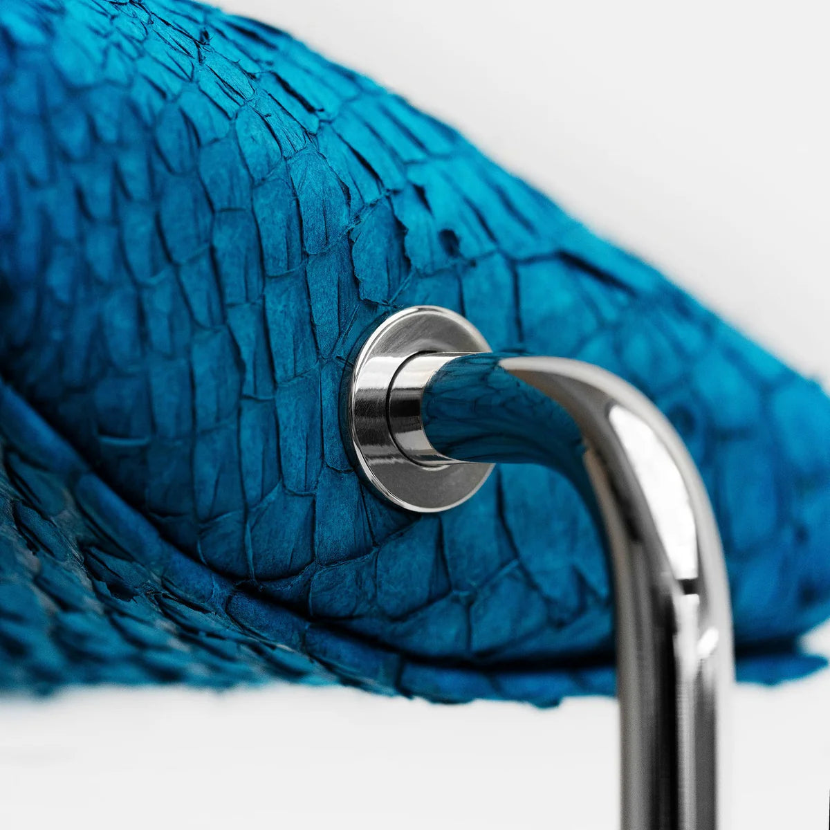 A close-up view of a blue textured cushion attached to a shiny silver curved metal support. The cushion has a scaly, reptile-like texture, and the metal support is secured with a polished silver screw. The image highlights the luxurious materials and the detailed craftsmanship of the display stand.