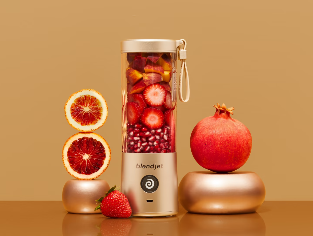 A stylish BlendJet blender filled with vibrant red and orange fruits like strawberries, pomegranate seeds, and sliced oranges, surrounded by fresh produce, including a whole pomegranate and strawberry, all set against a warm beige background.