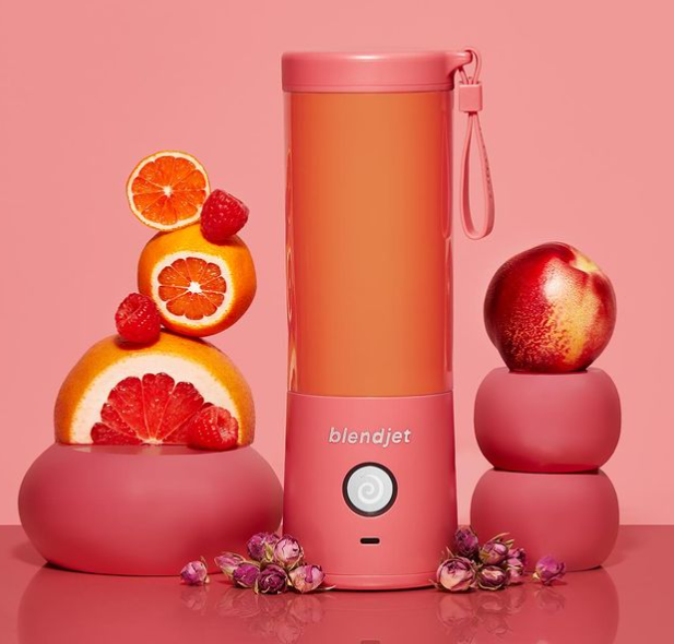 A vibrant coral BlendJet blender surrounded by stacked citrus fruits, raspberries, a nectarine, and dried rosebuds, all set against a pink backdrop for a lively and colorful presentation.