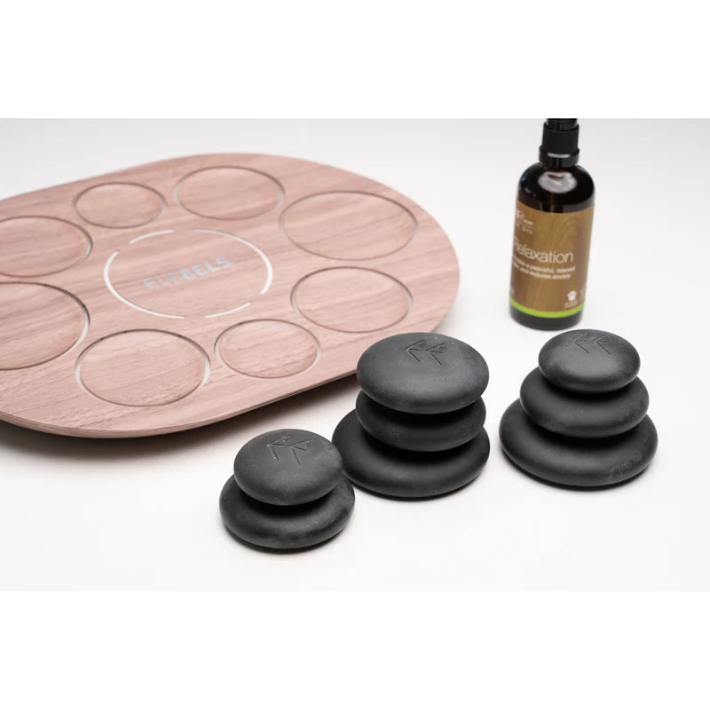 A complete ELEEELS hot stone therapy set featuring a wooden tray with recessed slots for organizing stones, stacks of smooth black hot stones engraved with the ELEEELS logo, and a bottle of ELEEELS Care Relaxation Massage Oil. The arrangement emphasizes the set's functionality and premium design for relaxation and therapeutic purposes.