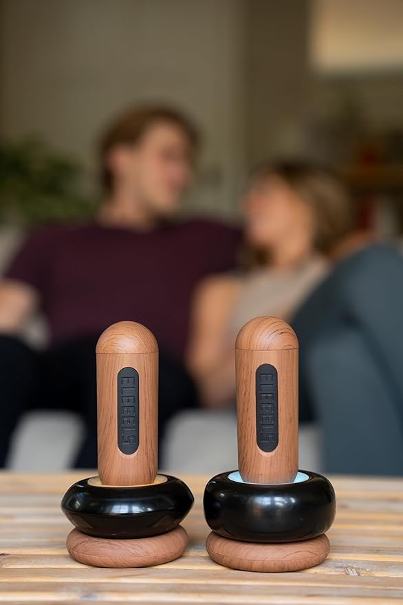 Two ELEEELS S2 Hot Stone Massage Wands with wooden handles and black bases are displayed on a wooden table, with a blurred background showing a relaxed couple sitting on a couch, highlighting the product's soothing and versatile use in a cozy setting.