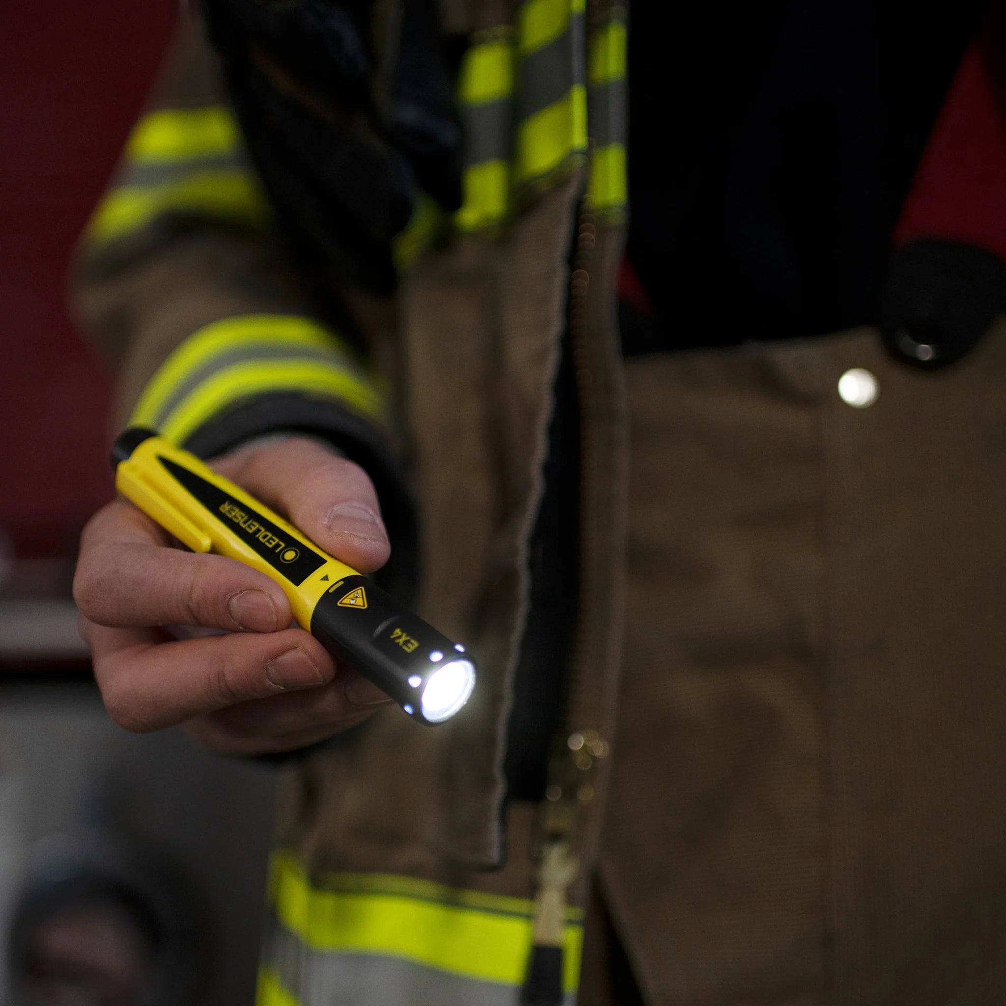 : A person wearing a firefighter's uniform is holding a lit LED Lenser EX4 penlight. The penlight's beam is clearly visible, illuminating the scene. The yellow and black design of the penlight contrasts with the uniform, highlighting its compact and portable design.