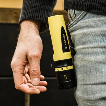 A person holding the LEDLenser EX7 flashlight against their side, with the flashlight tucked into the front pocket of their jeans. The yellow and black flashlight contrasts against the grey jeans and dark sweater.