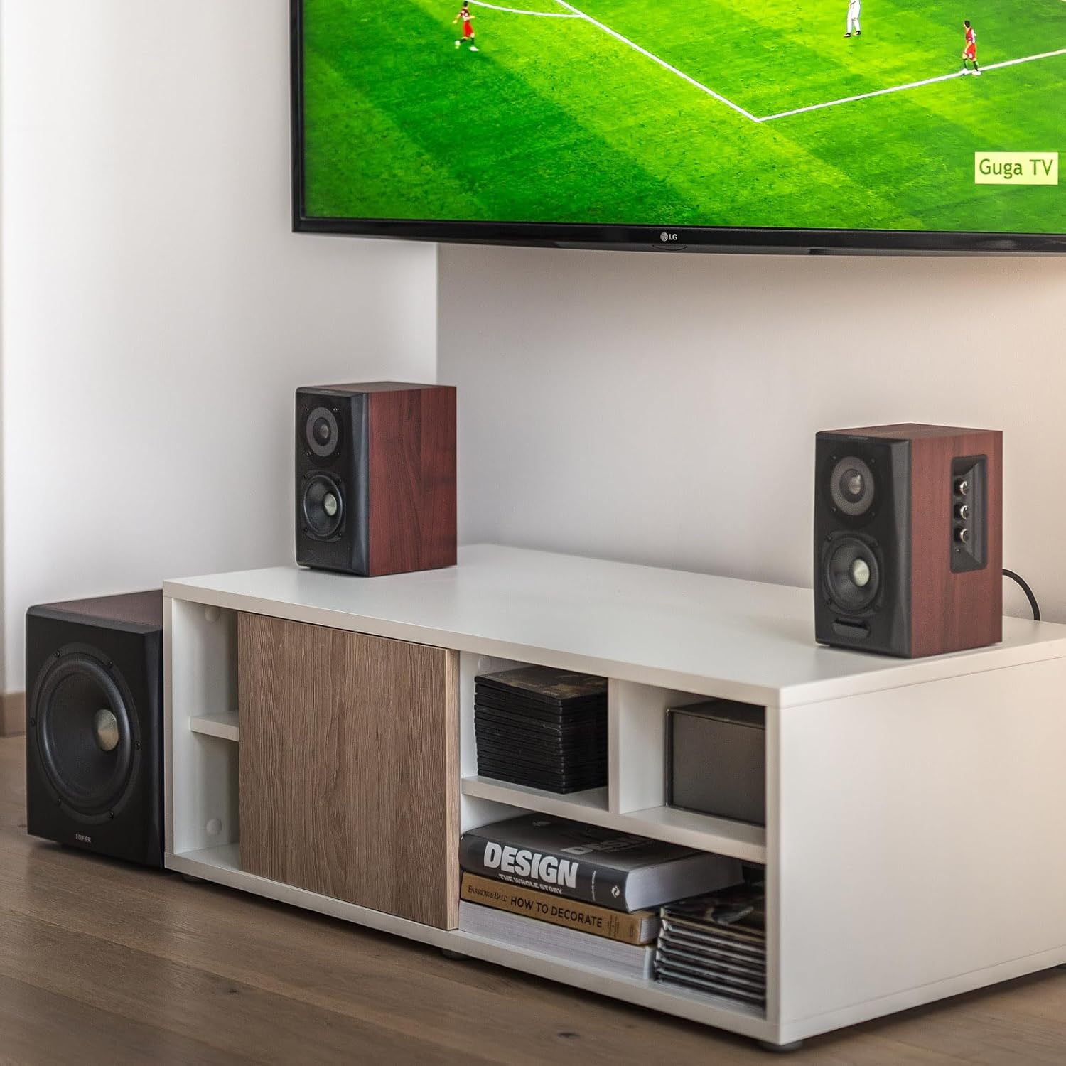 A modern living room entertainment setup featuring a flat-screen TV mounted on the wall, displaying a soccer match. Below the TV, a sleek white media console holds a pair of wooden bookshelf speakers with black front panels, positioned symmetrically. To the left of the console, a subwoofer is placed on the floor. The console also contains neatly arranged books and storage compartments, adding to the clean and organized aesthetic. The room features a minimalist design with a wooden floor and neutral-colored 