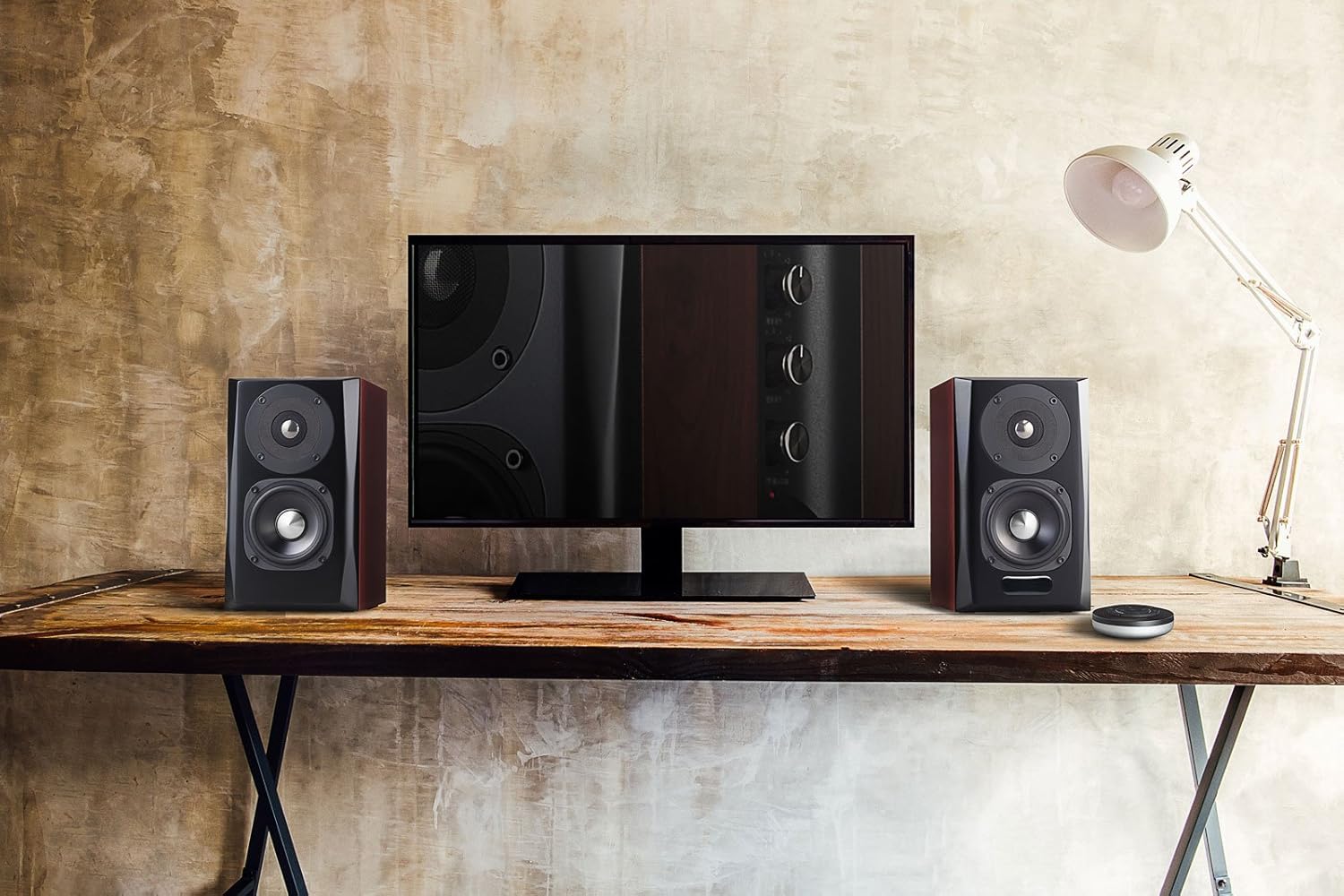 A stylish desktop audio setup featuring a pair of bookshelf speakers with a dark wood finish and a glossy black face. The speakers are placed on either side of a flat-screen monitor, which reflects part of the speaker's control panel. The setup sits on a rustic wooden table with a minimalist, industrial-style design. A small, modern desk lamp is positioned on the right, casting light onto the textured beige wall in the background, adding to the room's warm and cozy ambiance.