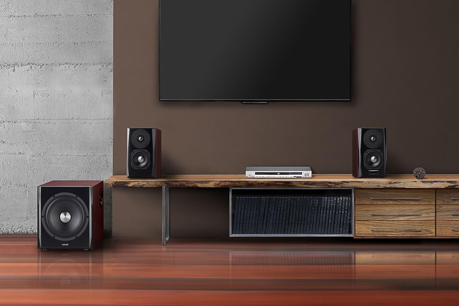 A modern home entertainment setup featuring a pair of bookshelf speakers and a subwoofer, all with a dark wood finish and black speaker grilles. The speakers are placed on a rustic wooden shelf beneath a wall-mounted flat-screen TV. A media player is positioned between the speakers, and a storage unit with books and drawers is built into the shelf. The room has a contemporary aesthetic with a mix of industrial and natural design elements, including a concrete wall and wooden flooring.