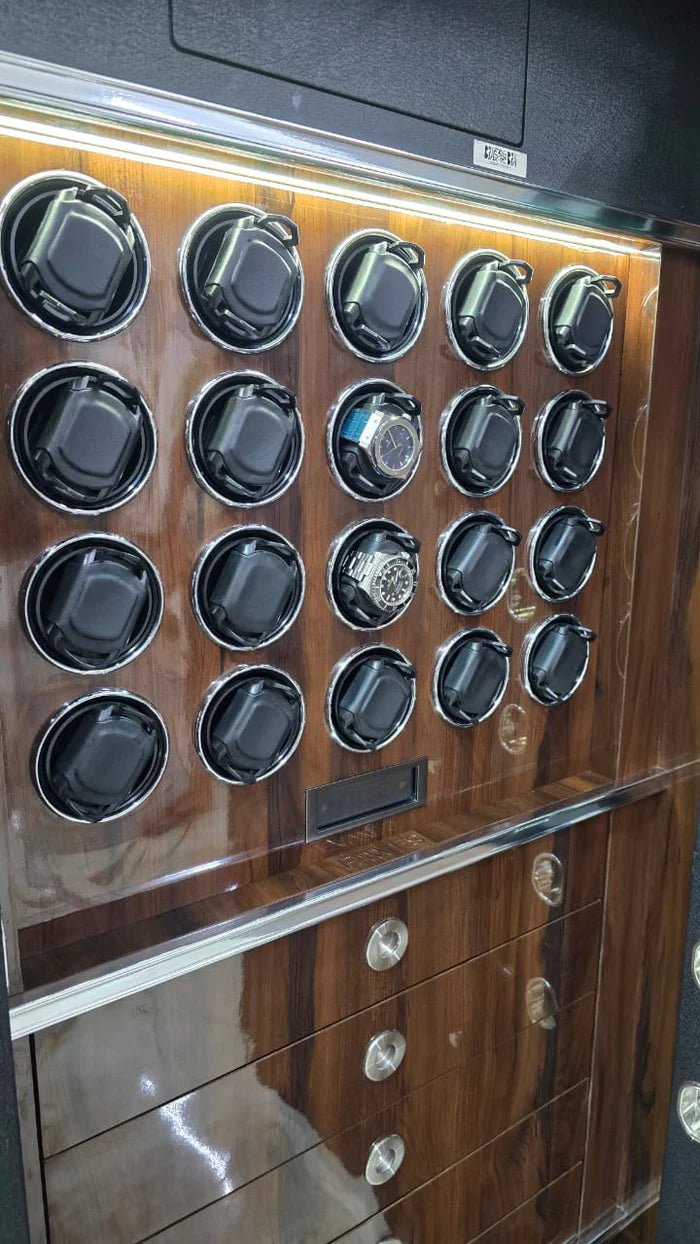 A close-up view of the interior of a large safe reveals multiple watch winders arranged in a grid on a wooden back panel. Some winders contain watches, while others are empty. Below the winders, there are three drawers with sleek handles for additional storage. The interior is illuminated with built-in lighting.