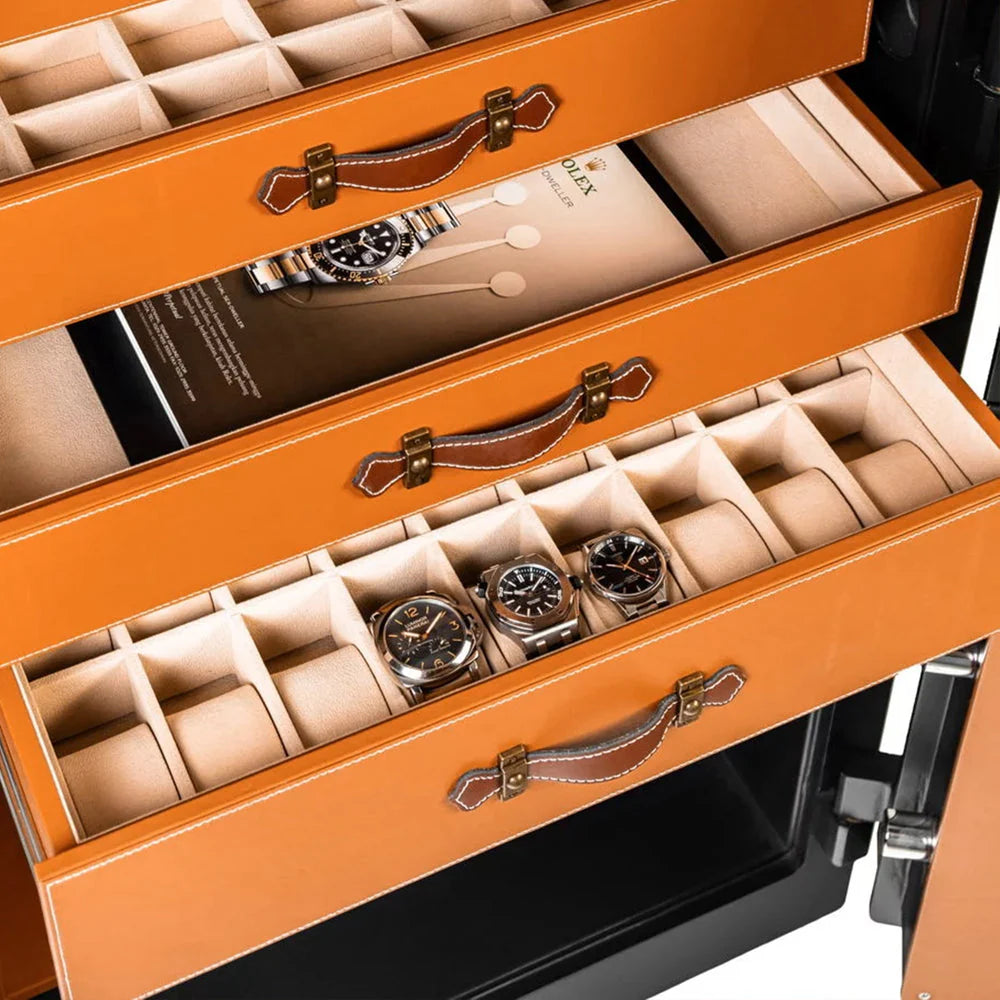 Close-up view of three open drawers inside a safe, each with an orange leather finish and elegant handles. The drawers are designed for organized storage of watches, with compartments lined with soft material to securely hold each watch. The top drawer holds a booklet, while the other two drawers display various luxury watches in individual compartments.