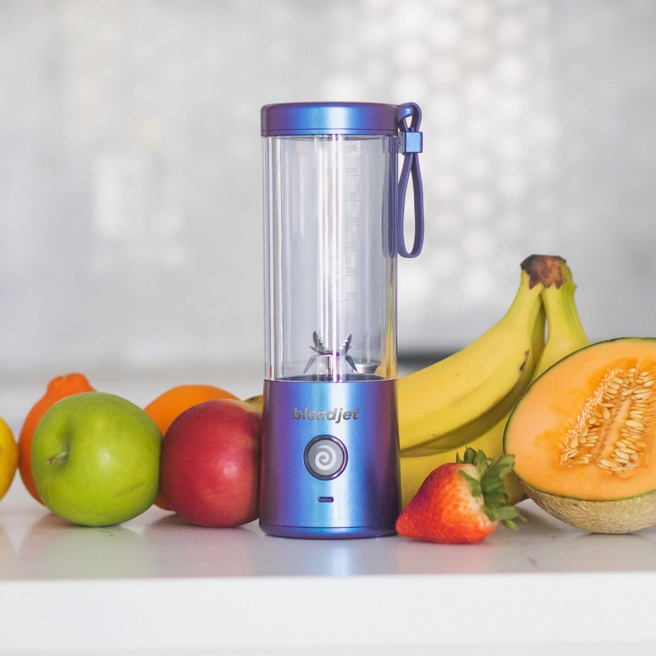 A compact blue portable blender with a transparent blending jar placed on a white countertop, surrounded by fresh fruits including a green apple, orange, red apple, bananas, half a cantaloupe, and a strawberry. The blender features a circular power button with a swirl design and a carrying strap attached to the lid. The background is softly blurred.