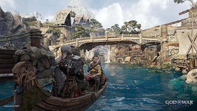 This image features a captivating scene from *God of War: Ragnarök*, showing Kratos and Atreus in a boat, journeying through a vibrant and lush village built along a serene waterway. The detailed setting highlights the architectural beauty of wooden structures and bridges, nestled against a mountainous backdrop. The water sparkles vividly under the natural light, with greenery and intricate carvings adding to the rich, mythological ambiance of the game.