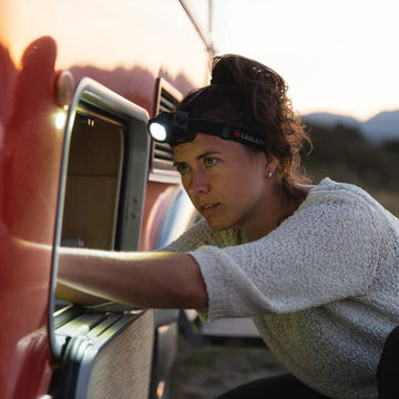 A woman wearing a LED Lenser headlamp is working on an outdoor task at dusk. She is focused and appears to be fixing or inspecting something on a red vehicle or structure. The headlamp illuminates her work area, highlighting its practical use in low-light conditions. The background shows a natural setting with hills and a sunset sky.