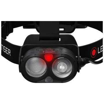 A close-up view of a LED Lenser headlamp showing the front light unit with two clear lenses and a small red indicator light in the center. The headlamp is attached to an adjustable black strap with the "LED LENSER" branding. The design emphasizes the dual-lens configuration for enhanced illumination