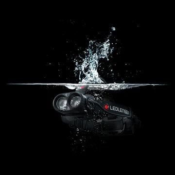  A black LED Lenser headlamp partially submerged in water, with a splash effect captured at the water's surface. The image emphasizes the headlamp's water-resistant or waterproof capabilities. The LED Lenser logo is visible on the headlamp's strap. The background is completely black, highlighting the splash and the headlamp.