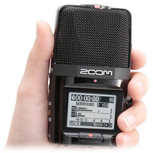 A hand holding the Zoom H2n Handy Recorder, showcasing its front microphone grille and large LCD screen displaying recording information and audio levels