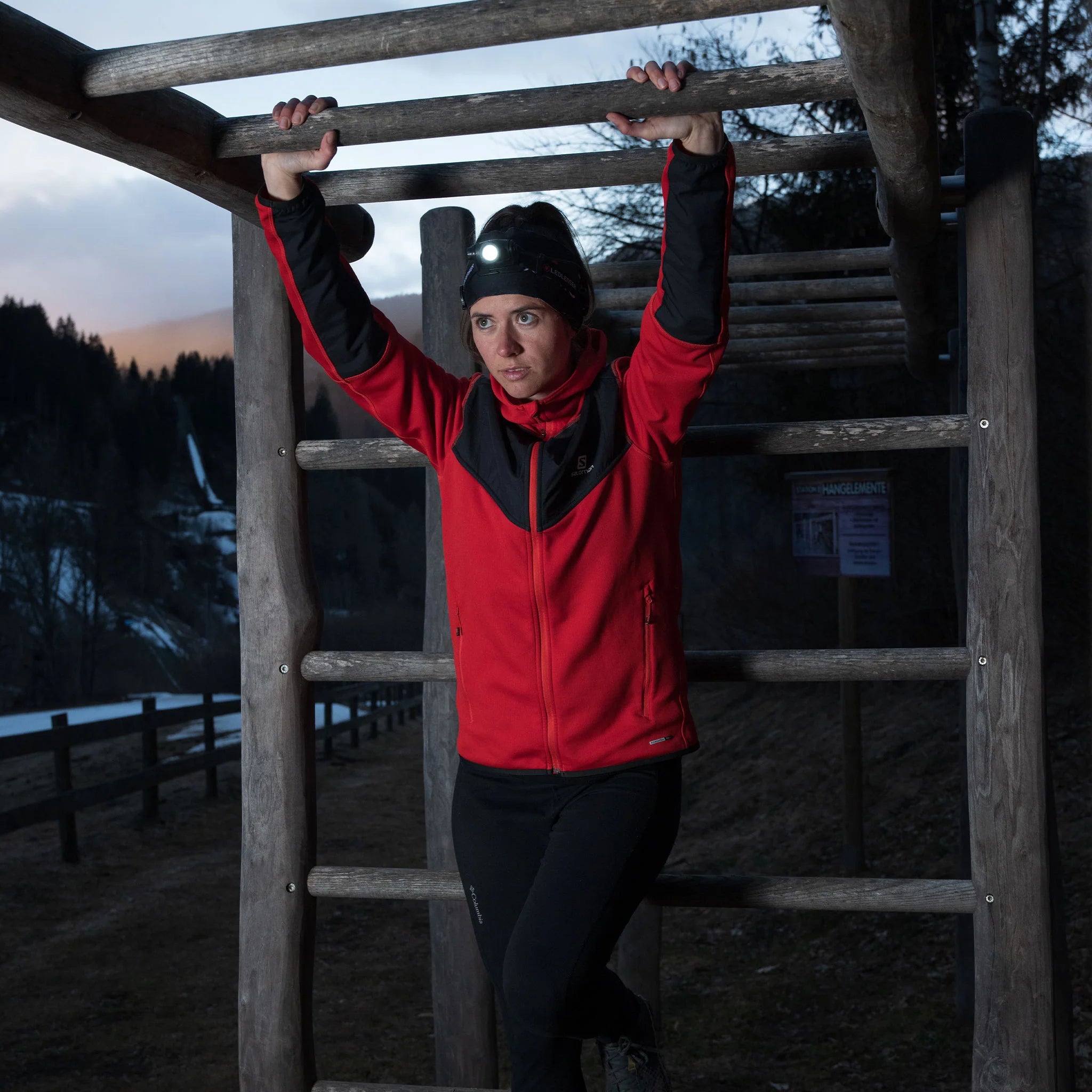  A woman wearing a red jacket and a black beanie with a LED Lenser headlamp is using an outdoor monkey bar structure at dusk. The headlamp is turned on, illuminating her face and the surrounding area. She is hanging from the bars, with a determined expression, in a wooded, mountainous area with a wooden fence and a sign visible in the background.