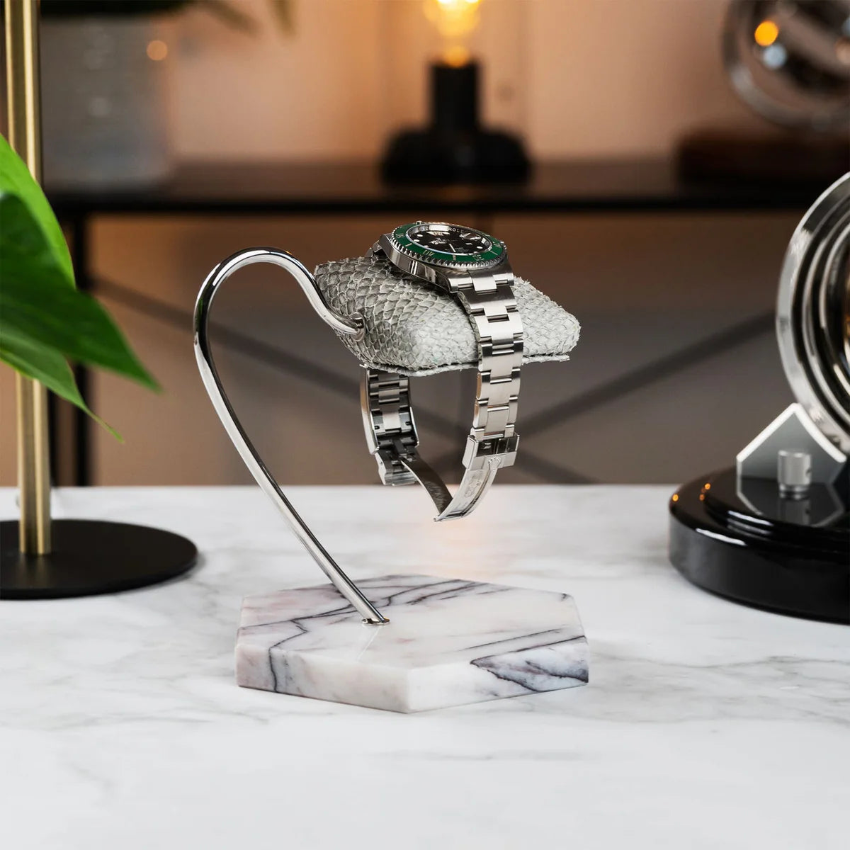  A wristwatch with a silver metal band and a green bezel is displayed on a textured gray cushion. The cushion is mounted on a sleek, curved metal stand with a hexagonal white marble base featuring gray veining. The setup is placed on a white marble surface, with a modern interior background that includes decorative elements like a plant, a gold lamp, and another watch display. The ambiance is elegant and sophisticated, highlighting the luxurious presentation of the watch.