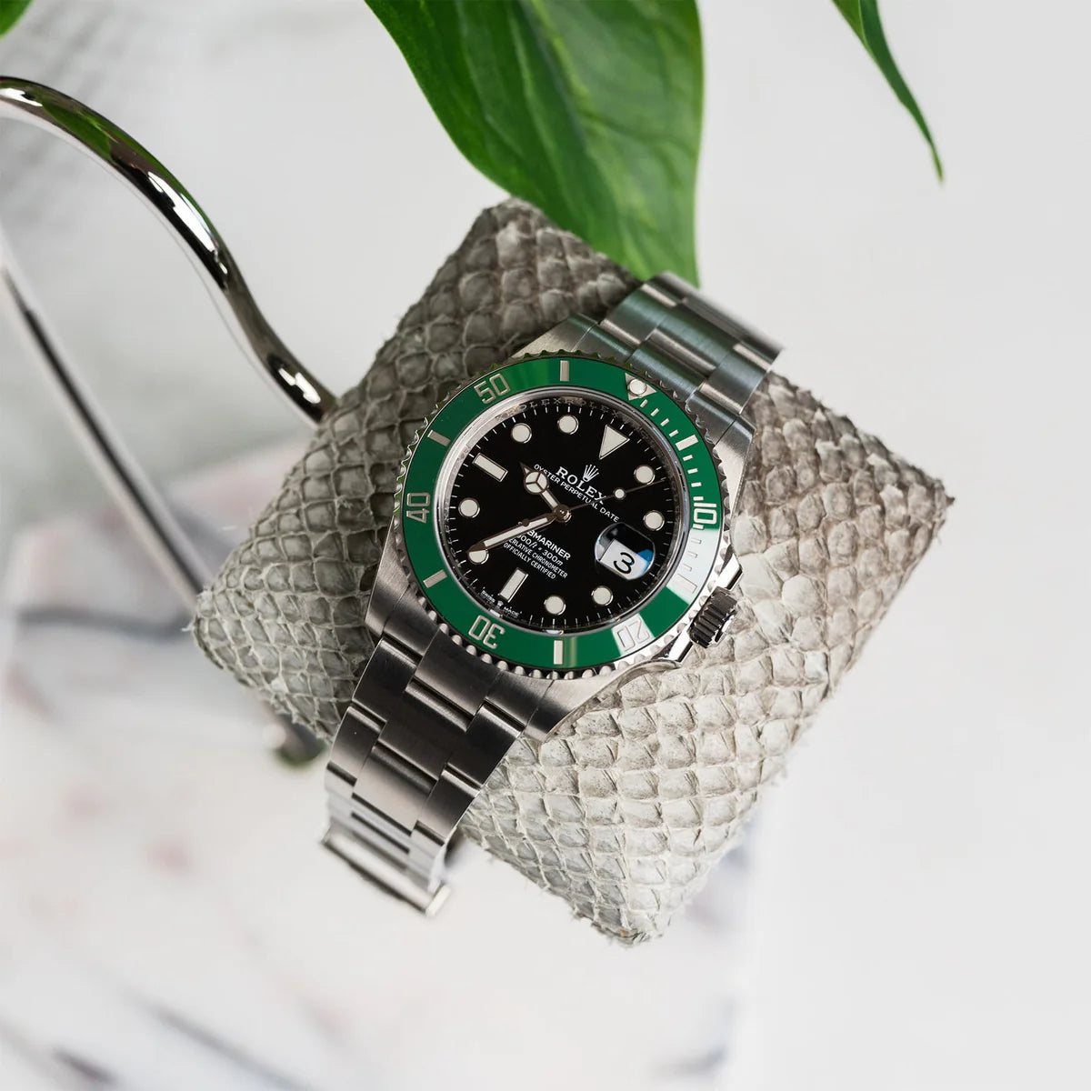  A stylish wristwatch with a silver metal band and a black dial with a green bezel is displayed on a textured gray cushion. The cushion is attached to a curved metal stand, which is partially visible. The background includes a green plant leaf, adding a natural element to the sophisticated and modern display setting.