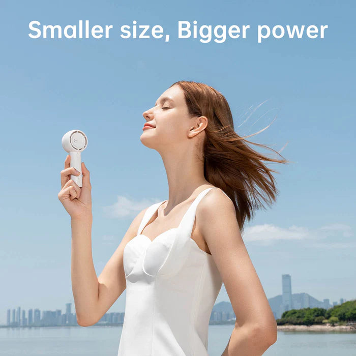 A woman standing outdoors, enjoying the breeze from a small, yet powerful handheld fan, showcasing the product's slogan "Smaller size, Bigger power" against a clear sky backdrop with a cityscape in the distance.