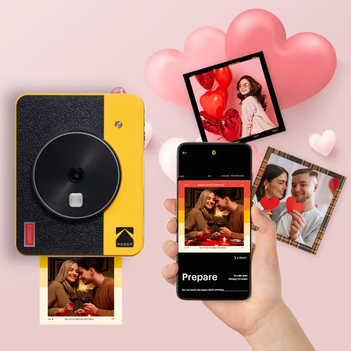 A Kodak instant photo printer in yellow and black is shown printing a picture, with a smartphone displaying the Kodak app interface for printing preparation, surrounded by printed images of couples and heart decorations, emphasizing easy and creative photo printing.