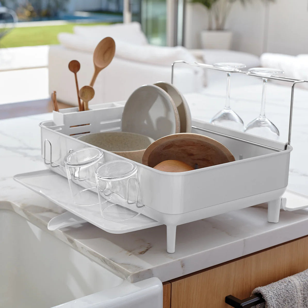 The image showcases a sleek white dish drying rack placed on a kitchen countertop. The rack is neatly organized, holding various kitchen items, including plates, bowls, utensils, and glasses. The design includes dedicated slots for plates, a utensil holder, and a special section for wine glasses, which are securely held upside down for drying. The rack is elevated on sturdy legs and features a side tray for additional items, with a draining system underneath to channel water away, keeping the countertop cle