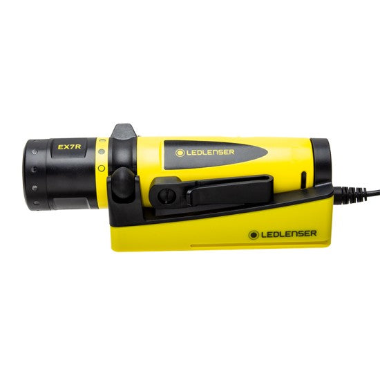 A yellow and black LEDLenser flashlight, model EX7R, placed horizontally in a matching yellow docking station for charging. The flashlight has a black head and a yellow body with the LEDLenser logo and model name printed on it. A charging cable is connected to the docking station.