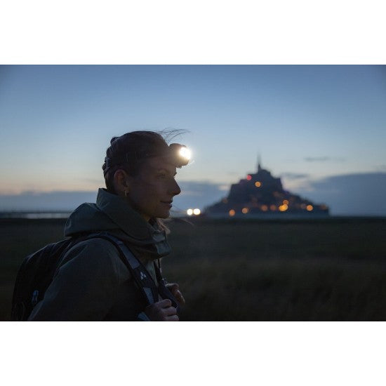 A woman wearing a headlamp during a dusk hike with a historical building illuminated in the background, showcasing the effectiveness of LED Lenser headlamps in outdoor and low-light environments.