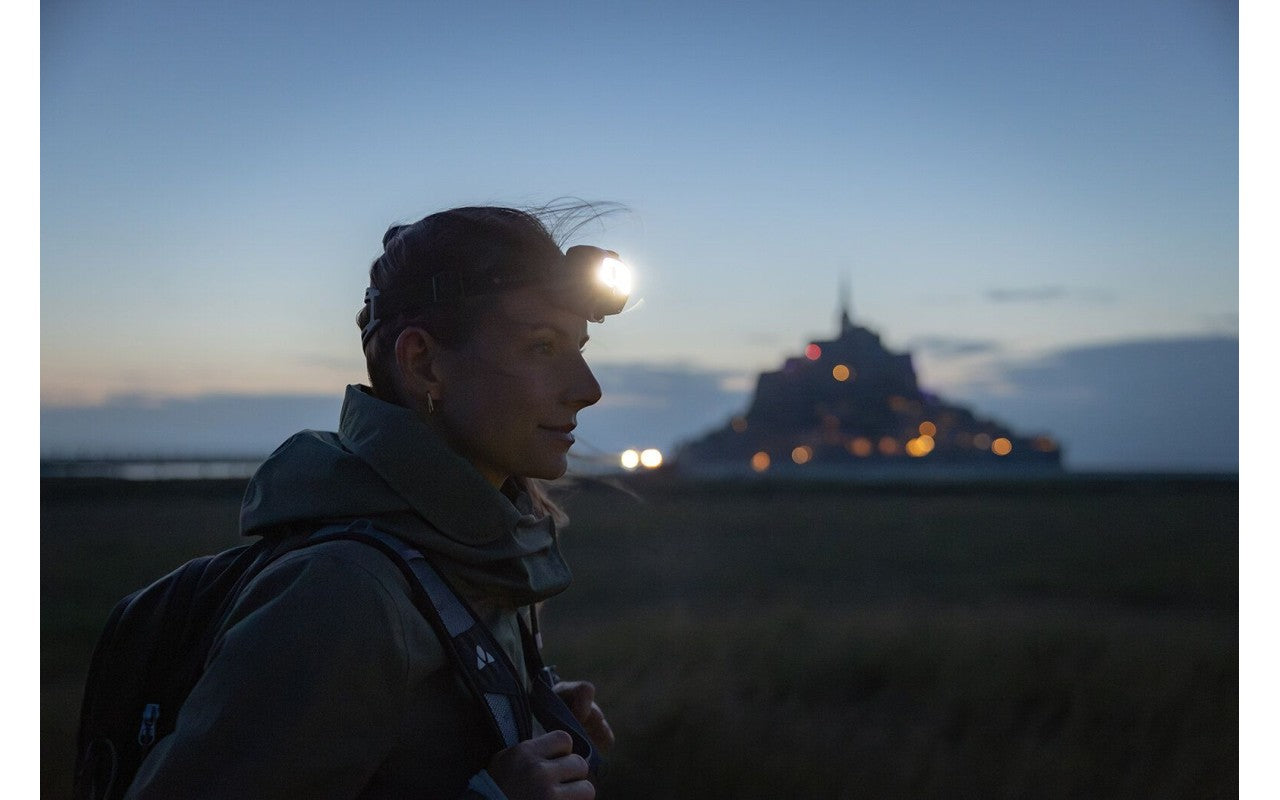 A woman wearing a LED Lenser headlamp, standing outdoors at dusk with a historic structure illuminated in the distance. The headlamp shines brightly, emphasizing its utility in low-light conditions. The serene evening sky and the soft glow of lights in the background create a peaceful yet adventurous atmosphere, capturing the essence of exploration and outdoor activity.