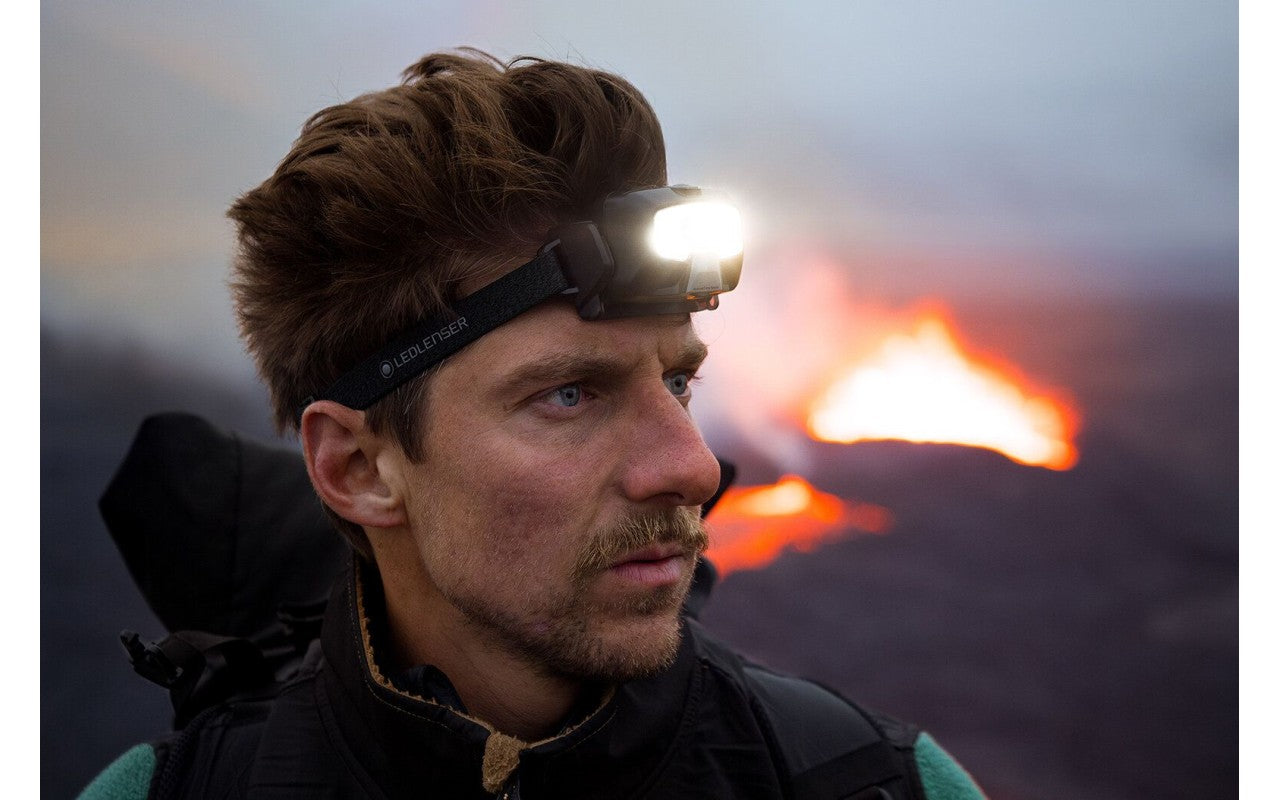 A man wearing a LED Lenser headlamp in a rugged outdoor environment, with a focused and determined expression. The bright headlamp illuminates the scene, highlighting the practicality of the device in challenging conditions. The background features an intense glow from what appears to be a distant fire or volcanic activity, contrasting with the cool tones of the setting.