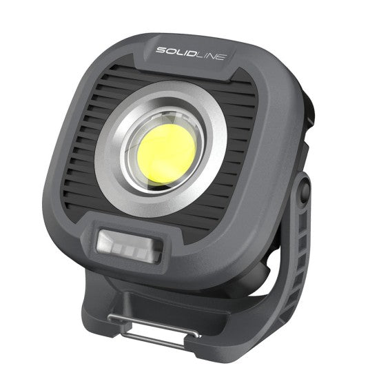  A compact, square-shaped black and gray LED work light from SOLIDline. The light features a large, circular LED in the center with a ridged design around it for heat dissipation. It has a sturdy base and a clip at the bottom for attaching or securing the light. The word "SOLIDline" is printed on the top edge of the light.