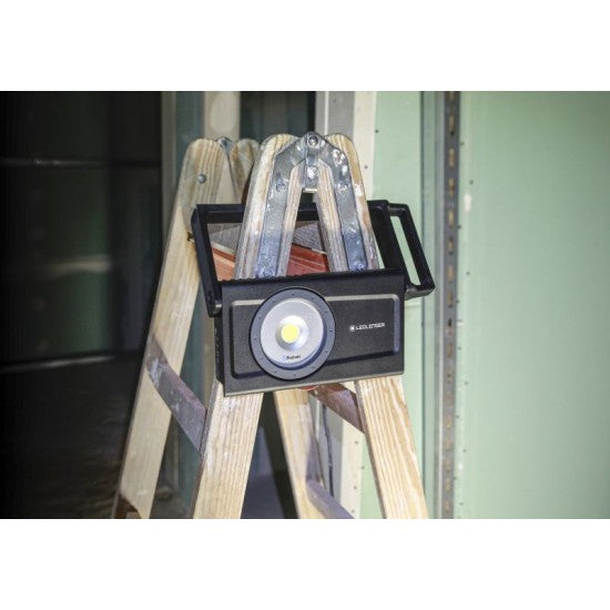  A LEDLenser portable light placed on top of a wooden ladder. The black light with a circular LED in the center is positioned to illuminate a workspace in a partially constructed or renovated room, with unfinished walls visible in the background.