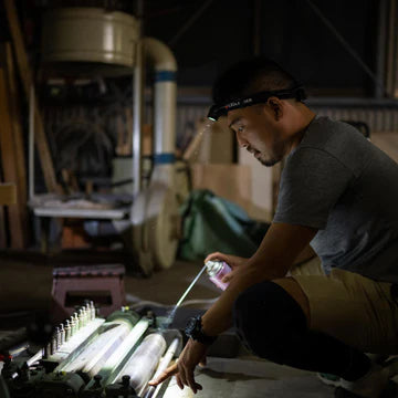 : A man wearing a LED Lenser headlamp is kneeling in a workshop, inspecting or repairing a piece of machinery. The headlamp illuminates his work area, allowing him to see clearly in the dimly lit environment. The workshop is filled with various tools and equipment, indicating a setting for detailed technical or mechanical work.