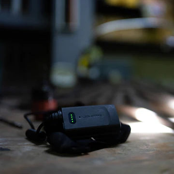 A close-up view of a LED Lenser headlamp's battery pack resting on a workbench. The battery pack has a green LED indicator showing the charge level. The background is blurred, with various tools and workshop equipment visible, indicating a setting for technical or mechanical work.