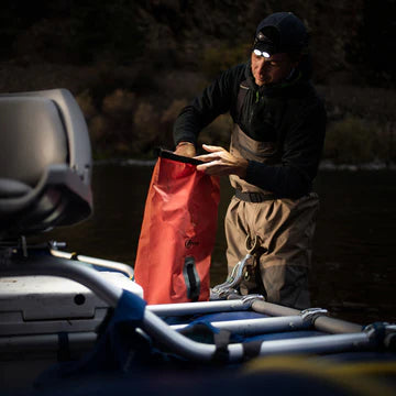 : A person wearing a headlamp is organizing a red waterproof bag on a boat at night. The individual is dressed in outdoor gear, including waders and a jacket, indicating a likely fishing or camping trip. The headlamp illuminates their work area, highlighting the practical use of the LED Lenser headlamp in low-light conditions. The background is dim, emphasizing the focused light from the headlamp.