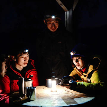  A group of four people wearing headlamps are gathered around a table at night, illuminated by a central lantern. They are engaged in conversation and appear to be in a relaxed, outdoor setting. The individuals are dressed warmly in jackets, indicating a cold environment. The scene is dark, with the headlamps and lantern providing the primary light source.