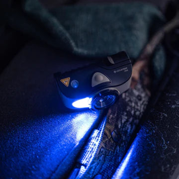 A black LED Lenser headlamp turned on, emitting a bright blue and white light. The headlamp is resting on a textured surface, illuminating the area around it. The light creates a strong beam, showcasing the headlamp's powerful illumination capabilities.