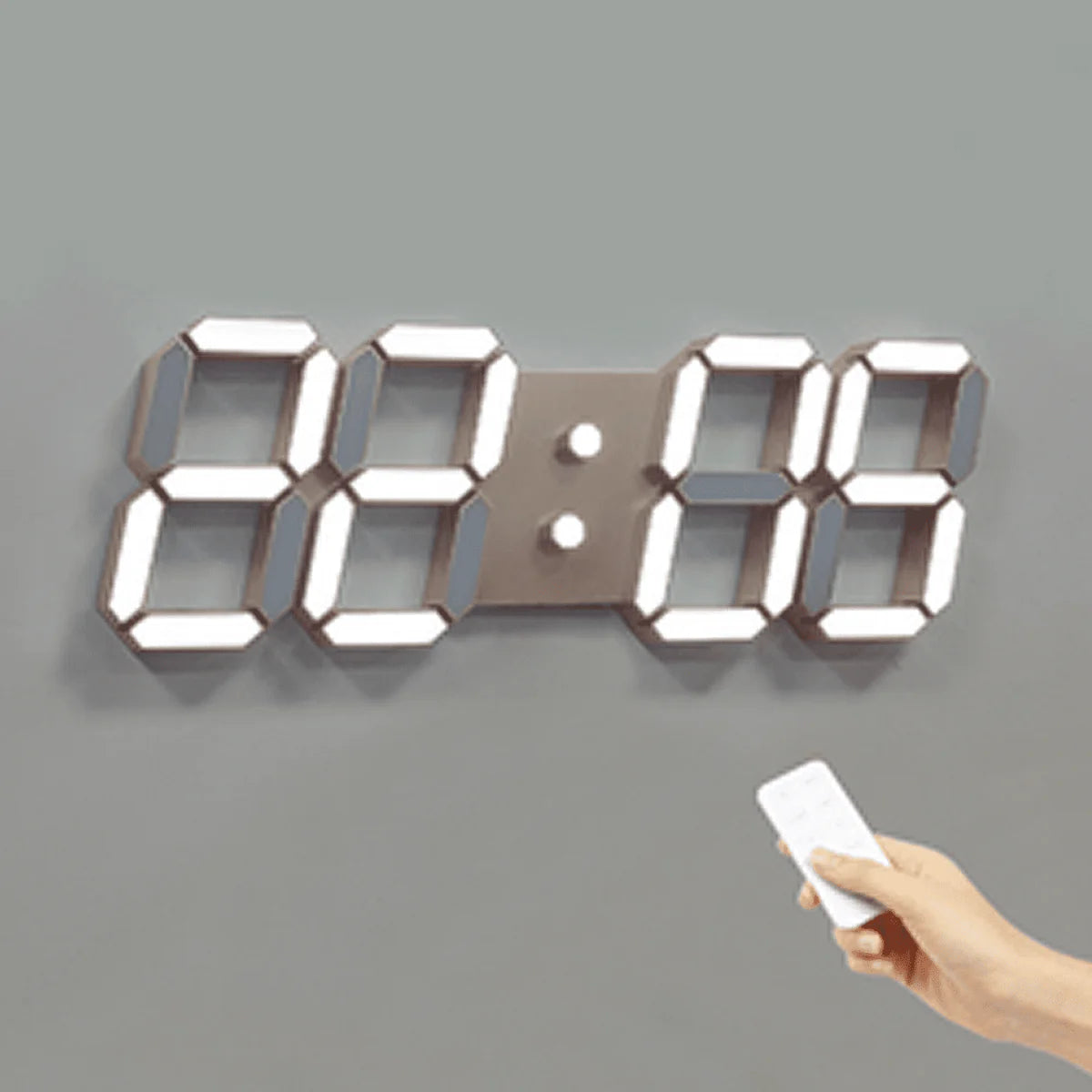 A digital wall clock with large illuminated numbers is mounted on a gray wall. The clock is displaying "88:88." A hand holding a remote control is aimed at the clock, indicating the use of the remote to set or adjust the clock. The clock has a sleek, modern design with bright, easily readable numbers.