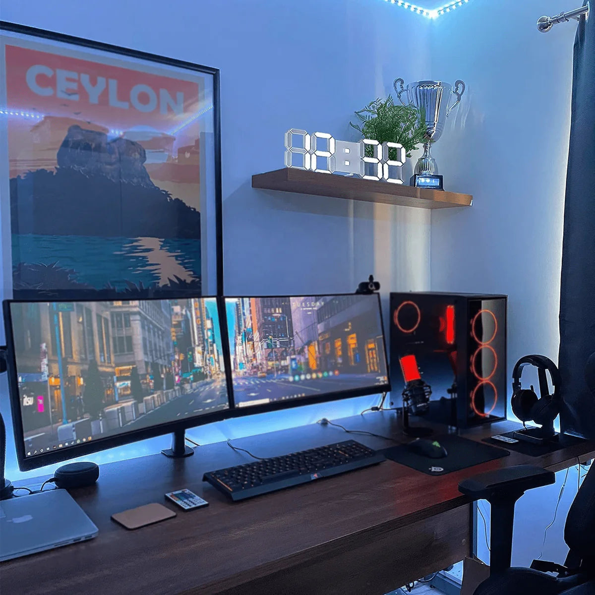  A modern gaming setup featuring dual widescreen monitors on a wooden desk. The room is illuminated with blue LED lights, enhancing the gaming ambiance. On the right side, a gaming PC with red interior lighting and a headset hanging on a stand is visible. Above the monitors, a shelf holds a digital clock and a trophy. To the left, a framed poster with "CEYLON" in bold letters adds a decorative touch to the setup