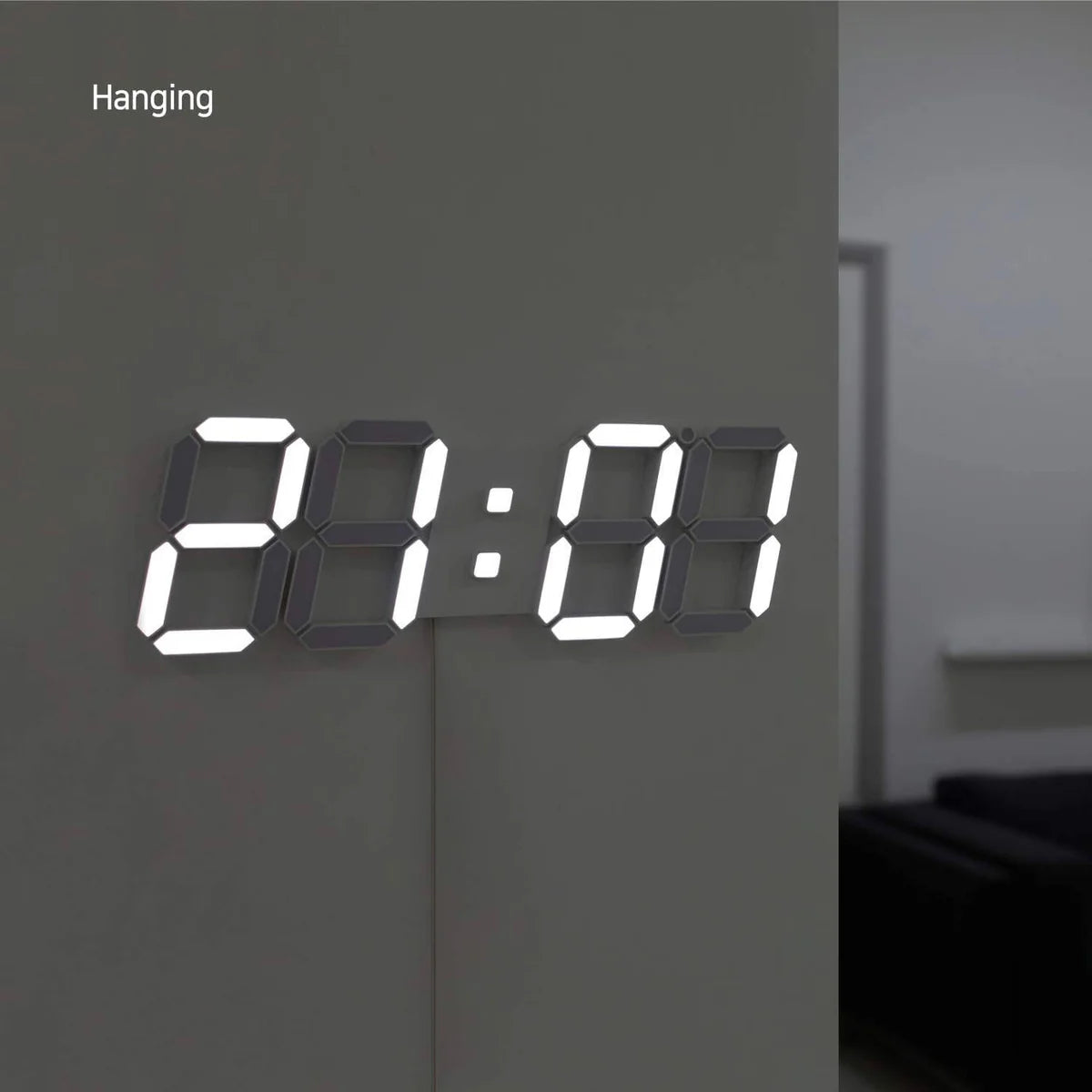  A digital wall clock with large illuminated numbers is mounted on a wall corner, displaying the time. The room is dimly lit, and the clock's bright display stands out against the neutral wall. The setting appears to be a modern interior with minimalist decor, featuring a glimpse of a sofa and a partially visible room in the background. The clock is labeled "Hanging," indicating its mounted position.