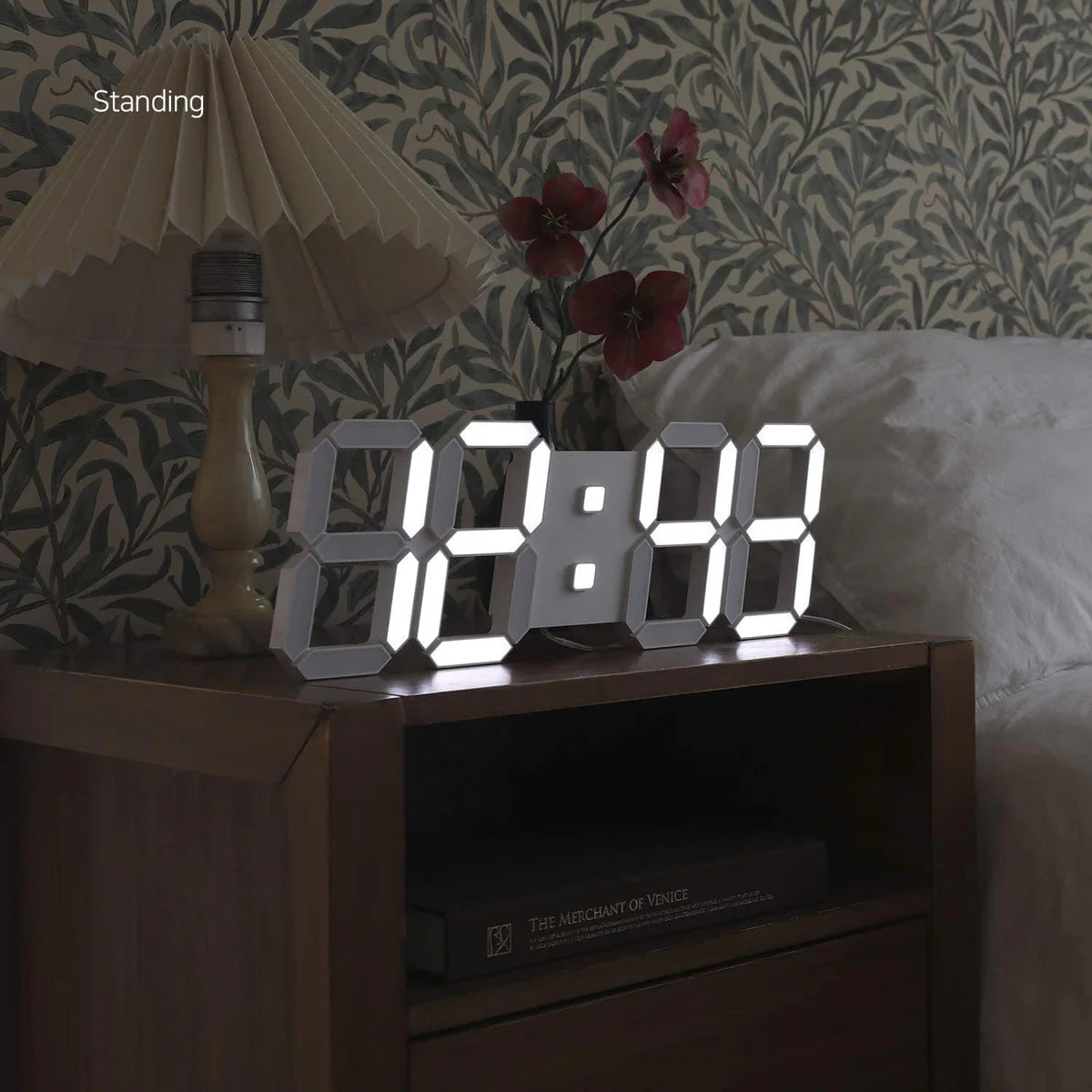A bedroom setup with a wooden nightstand next to a bed. On the nightstand, there is a digital clock with large illuminated numbers displaying the time. Next to the clock, there is a vintage-style lamp with a pleated lampshade and a few decorative red flowers. The wallpaper in the background features a floral pattern, adding a classic touch to the cozy and intimate setting.
