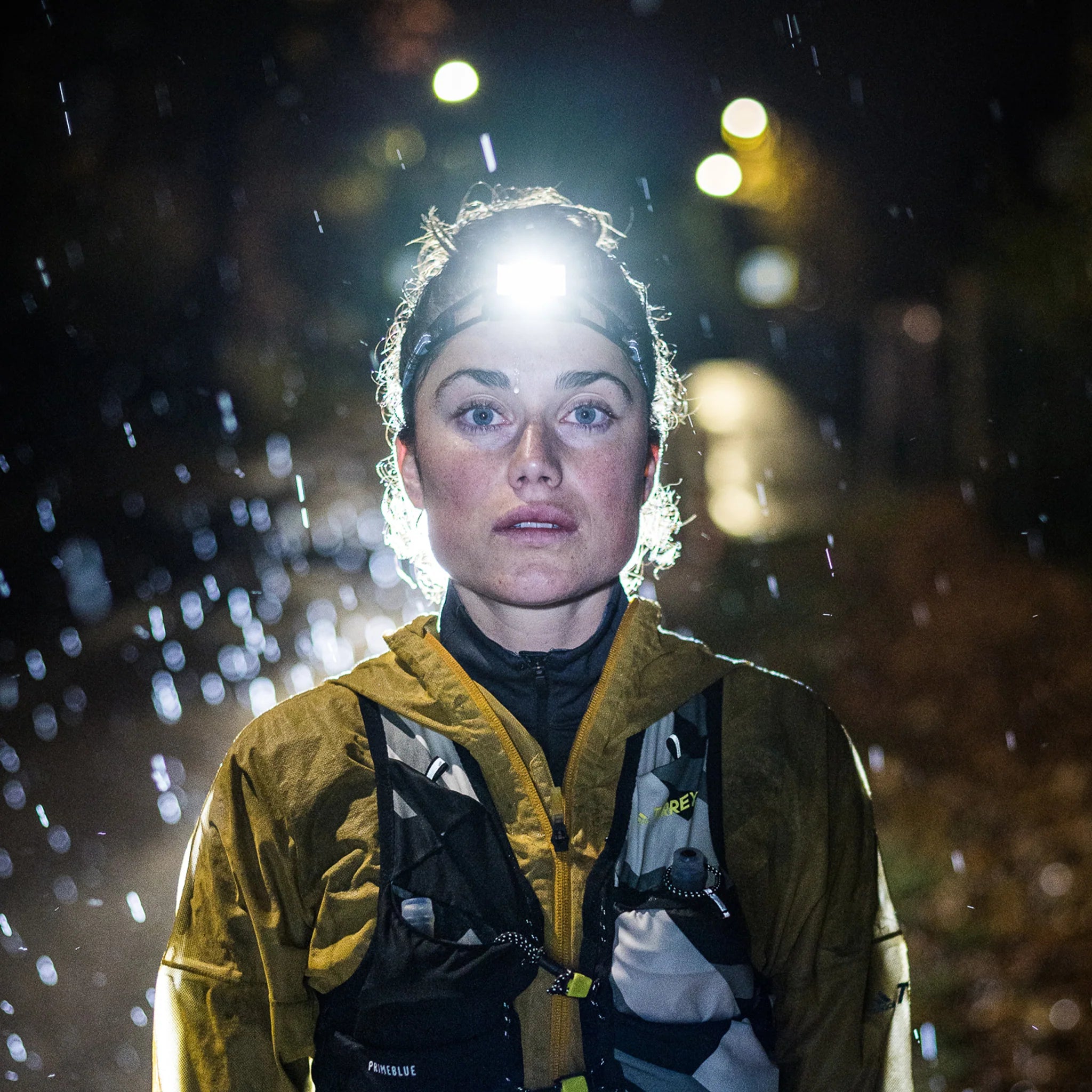 A woman wearing a headlamp and a yellow waterproof jacket is running at night in the rain. The headlamp is turned on, illuminating her face and the path ahead