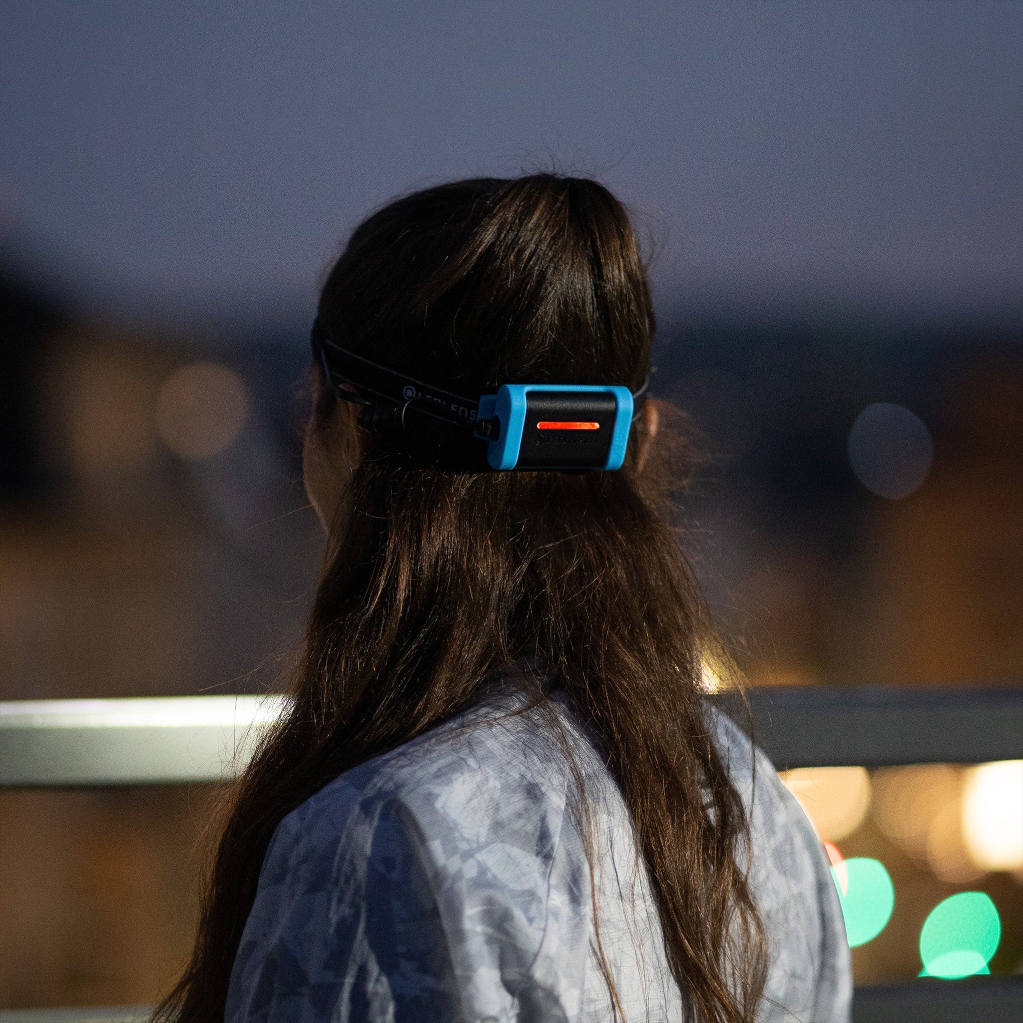 A person viewed from behind, wearing a headlamp with a blue and black strap, featuring a visible red light at the back. The scene is set during dusk or early night, with the background softly blurred, highlighting the headlamp's illumination.