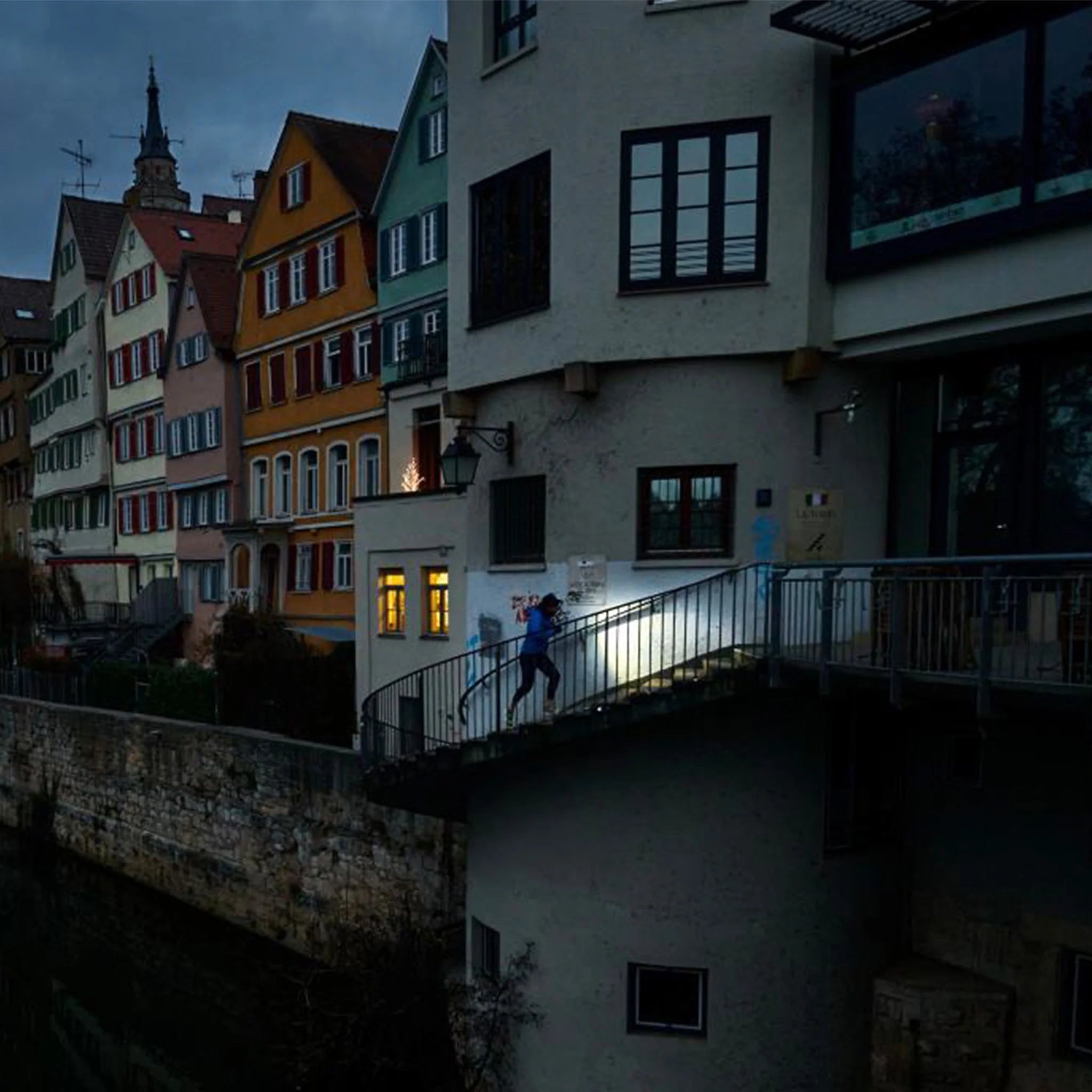 A person running up a stairway in the evening, illuminated by a bright light, with a background of colorful, traditional European buildings near a waterway. The scene captures a mix of urban and natural elements in a quaint town.